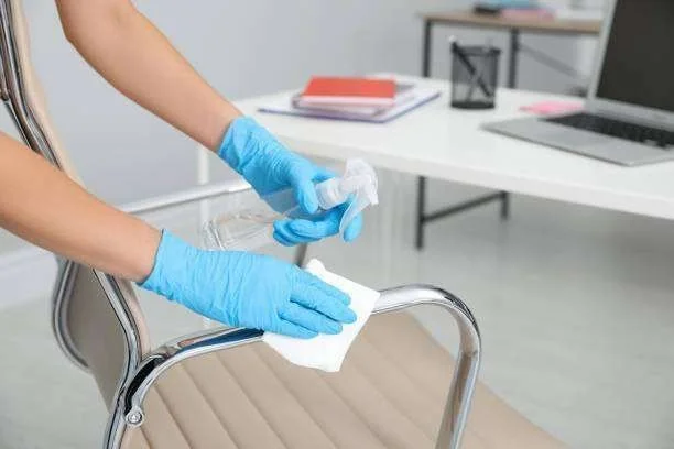 Mãos com luvas azuis higienizando o braço cromado de uma cadeira de escritório com spray e pano